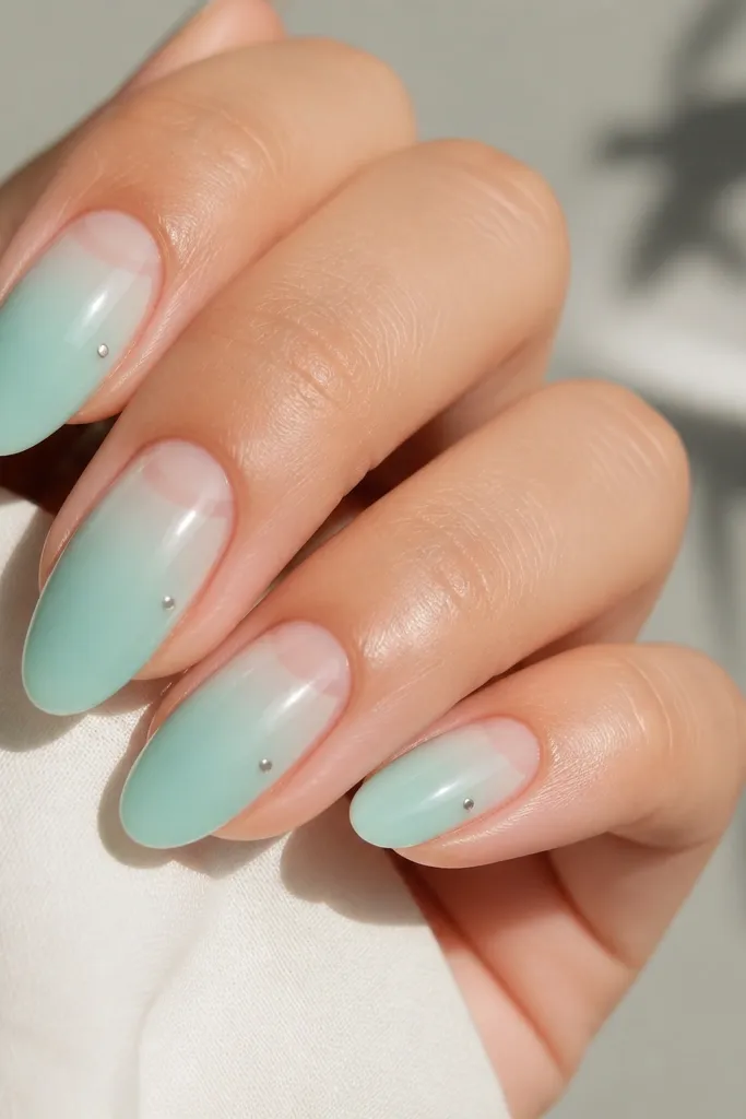 Four almond nails in a pale mint-to-aqua fade with a clear negative-space half-moon near the cuticle; the rest is glossy with a tiny silver dot on one side of each nail.