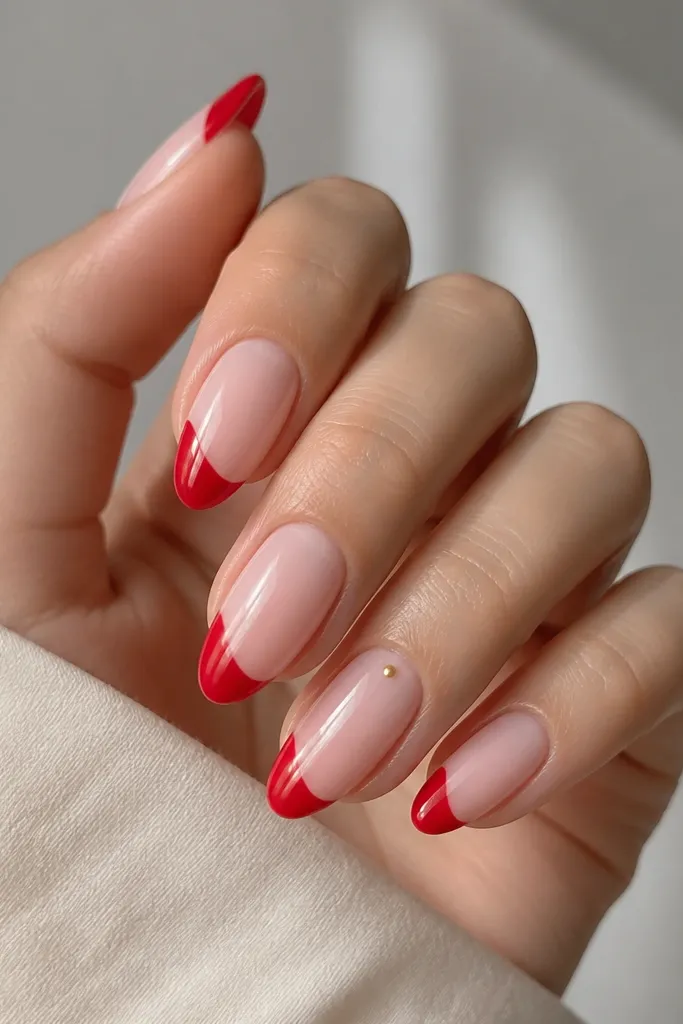 Almond nails with a milky pink base and glossy strawberry red tips shaped like rounded commas; one nail has a single tiny gold dot near the cuticle.