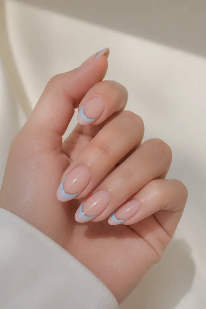 Natural almond nails with a nude base and baby blue half-moon at the tip side, leaving most of the tip clear. The line is thin and forms a smooth crescent shape.