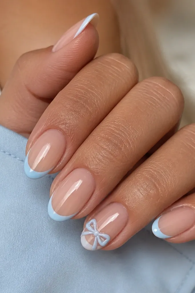 Nude base with baby blue French tips. On the ring finger, there is a small painted bow in baby blue and white near the smile curve, glossy and dimensional.