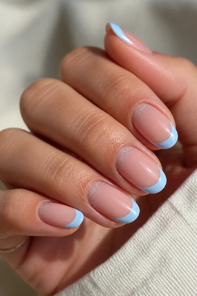 Nude base nails with baby blue French tips. Around the cuticle area there is a tiny halo of micro glitter, subtle and only visible close up. Glossy finish.
