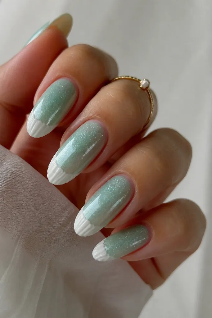 Long almond nails painted semi-sheer sea foam green with fine iridescent glitter. At the tip, a half-moon shell cap shape in white sits like a crescent.