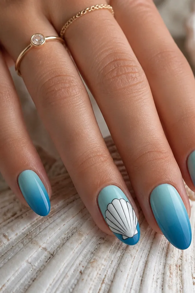 Medium almond nails with vertical ocean ombre: light aqua near the cuticle fading into deeper blue at the tip. On the ring finger, a large white shell spans most of the nail with tiny dark outlines.