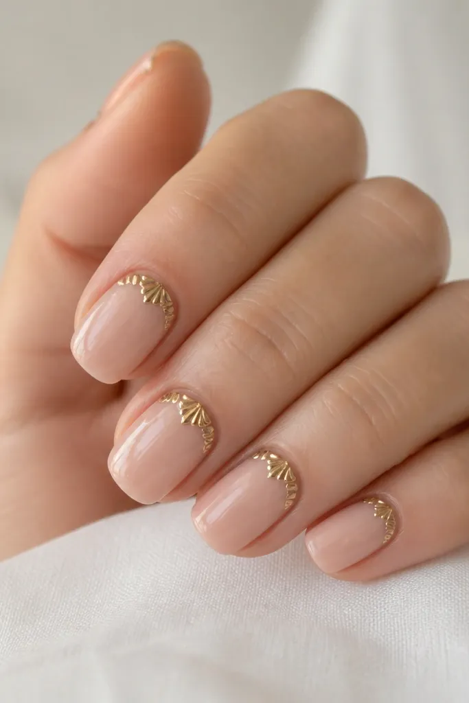 Short squoval nails with nude base and a glossy finish. At each cuticle, there's a small gold scallop crown made of shell-like shapes. The rest of the nail stays plain.