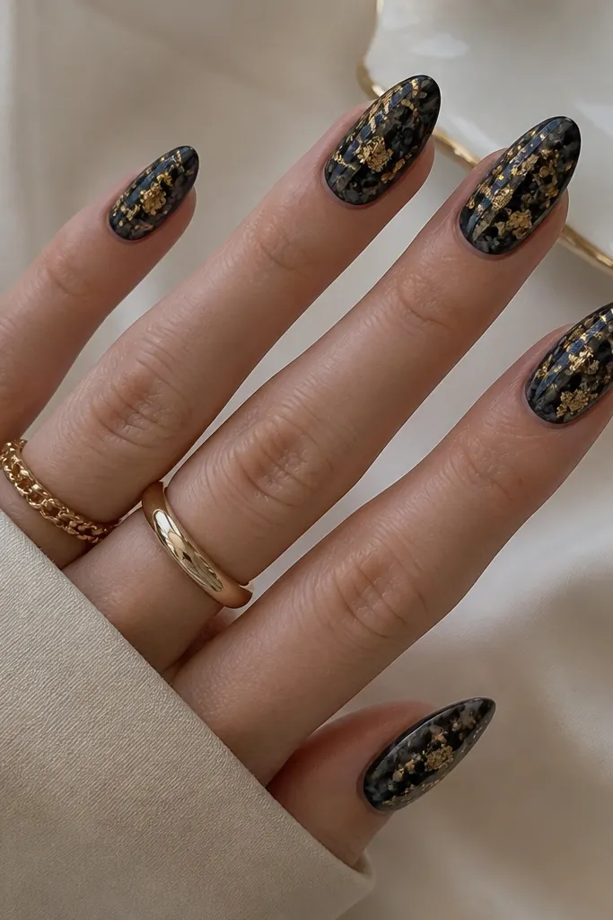 Almond nails with a smoky black marble base. Thin gray-black veins swirl throughout. Gold leaf flecks sit mostly around the center and near the tip, with a few pieces near the cuticle.
