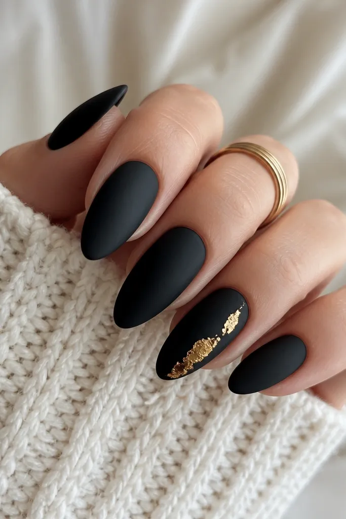 Almond nails with matte black base. On one accent nail, a warm yellow-gold foil drip starts near the center and falls toward the tip in two uneven streaks, with foil pieces overlapping like molten metal.