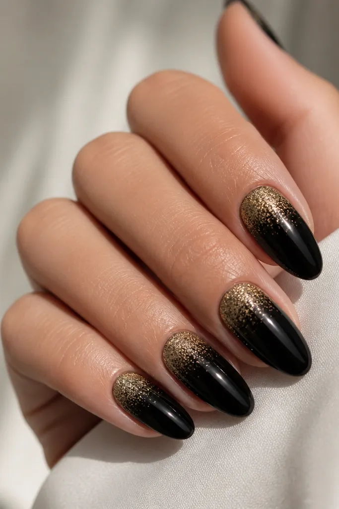 Almond nails with glossy black. At the cuticle, a gold micro-glitter fades downward into the black, stopping around the middle. The glitter is densest near the cuticle and sparse toward the fade edge.