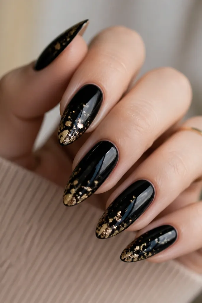 Almond nails with glossy black. Gold foil confetti specks scatter across the nail surface like tiny irregular flakes. The heaviest cluster sits near the tip on each nail, with a few specks near the middle.