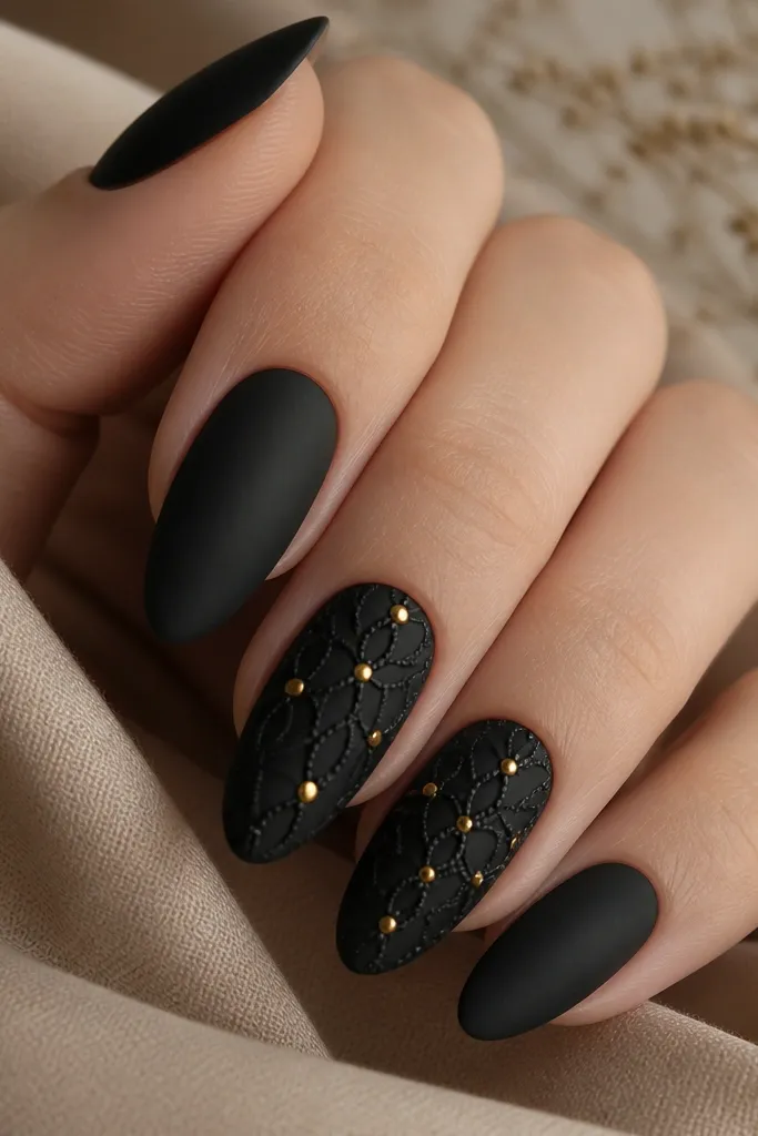 Almond nails with matte black base. Over two accent nails, there's a lace pattern made of thin raised lines and small loops in black, with warm gold dots at key intersections. The rest are solid matte black.
