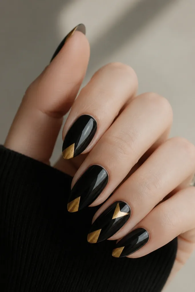 Almond nails with glossy black. At the tip, a geometric triangle is painted in warm gold, with crisp edges. One or two nails have a second smaller gold triangle near the side, still aligned to the tip geometry.