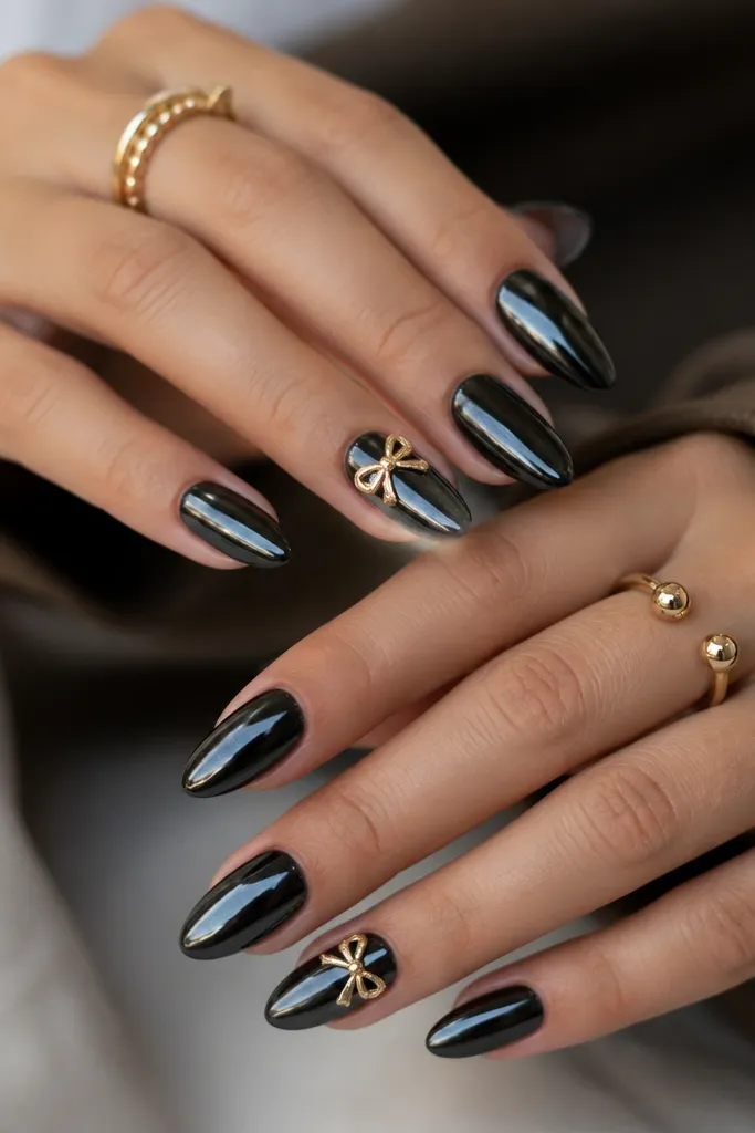 Black glossy chrome nails with one or two accent nails featuring a small gold ribbon bow at the cuticle area, bow tied with metallic sheen.