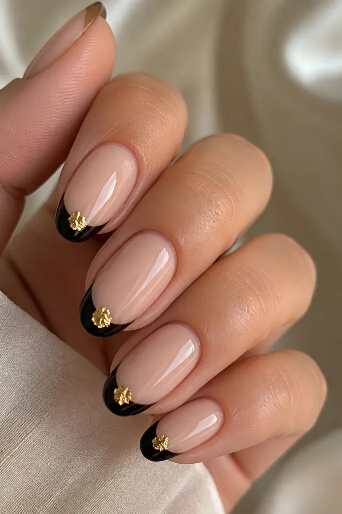 Black French tips on nude nails with a small gold leaf triangle centered at the very tip. The triangle has uneven leaf edges and catches light like foil fragments.