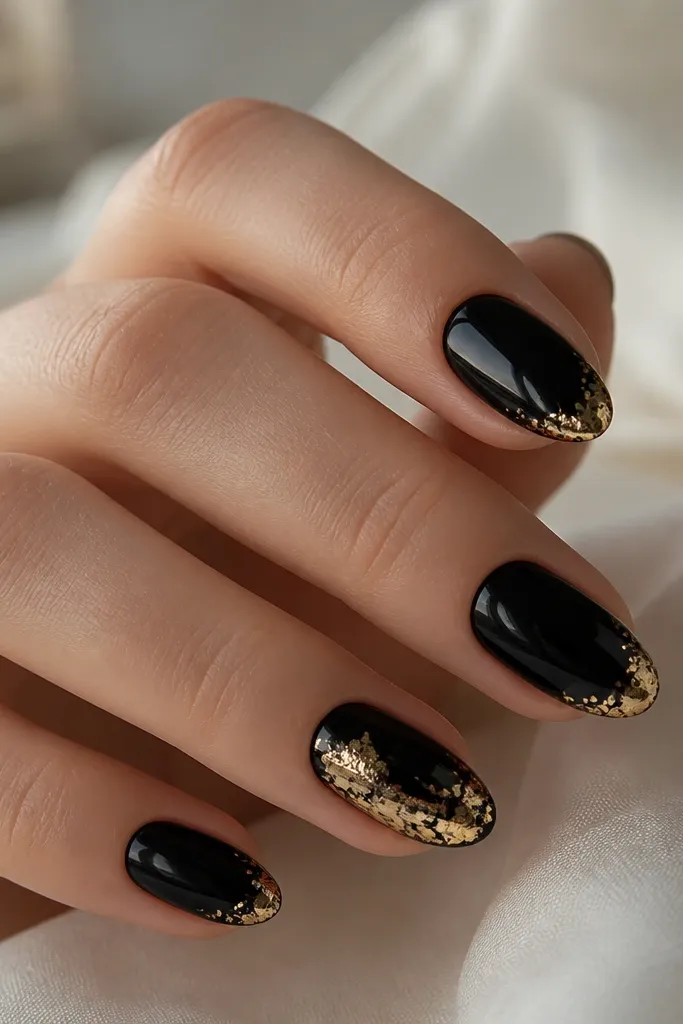 Short almond nails with a glossy black base. The tips have thin, irregular gold foil patches concentrated on the outer corners, with a few tiny foil flecks fading inward. One nail has slightly heavier foil coverage.