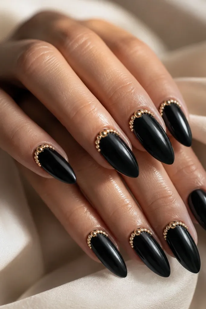 Long almond nails in glossy black. A crescent of small gold rhinestones hugs the cuticle on each nail, with stones getting slightly larger at the center. The rest stays plain black.