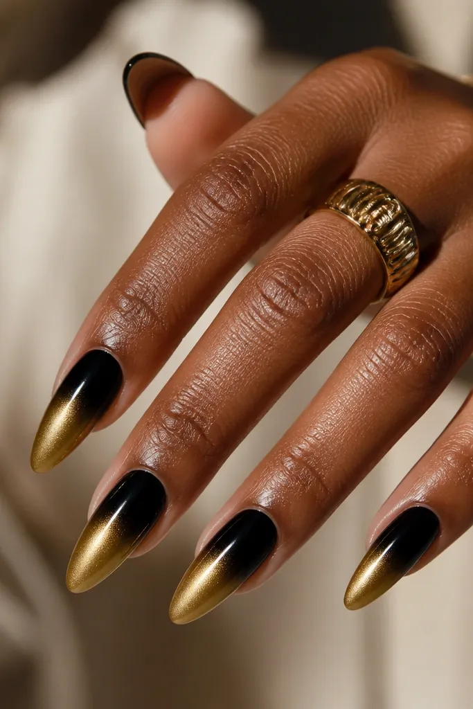 Long almond nails with an ombre that starts black at the cuticle and fades into gold near the tips. The gold area is a soft metallic gradient, not a harsh line. Everything is glossy.