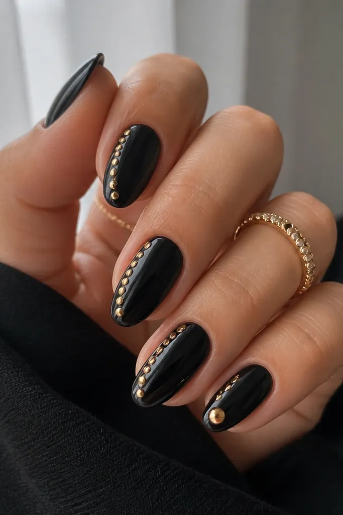 Medium oval nails in glossy black with a line of small gold studs along the outer sidewall of each nail. The studs stop around the mid-nail, tapering slightly. One accent nail has a single larger gold stud near the tip.