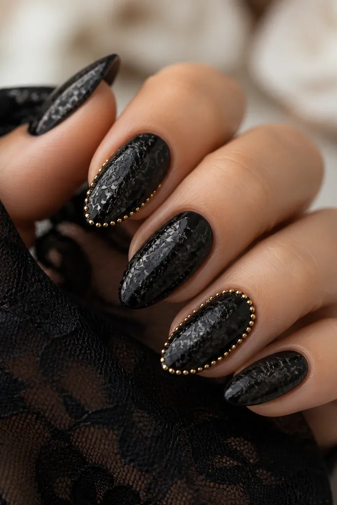 Nails painted glossy black. On top, a lace pattern is drawn in a slightly raised black or dark gray linework. Around the perimeter of two accent nails, there's a gold dot border tracing the nail edges.