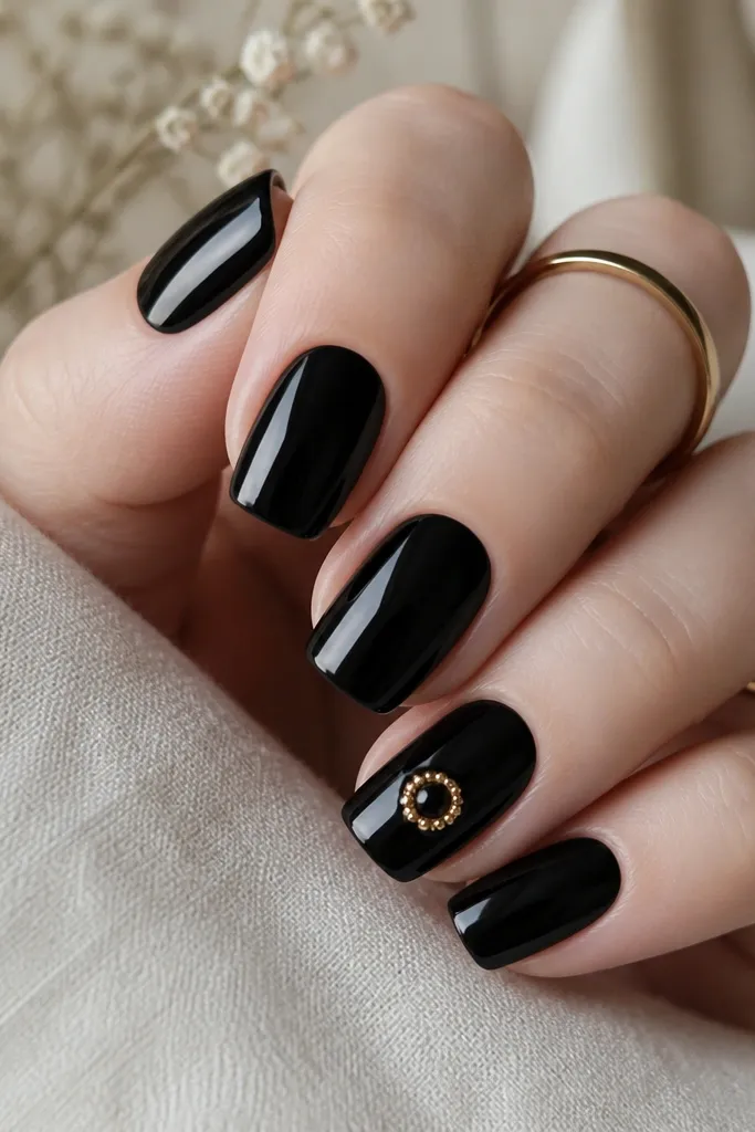 All nails glossy black except one accent nail. The accent nail has a gold rhinestone halo around a small central gem near the middle of the nail. The halo stones are tiny and evenly spaced.