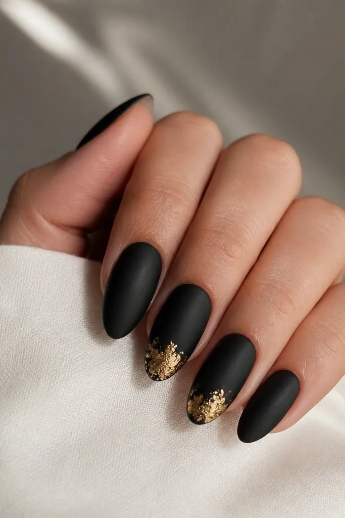 Medium almond nails with satin-black finish. At the tip on two accent nails, there's a clustered patch of gold leaf concentrated toward the center, with a few wisps spreading outward. Other nails are plain satin black.