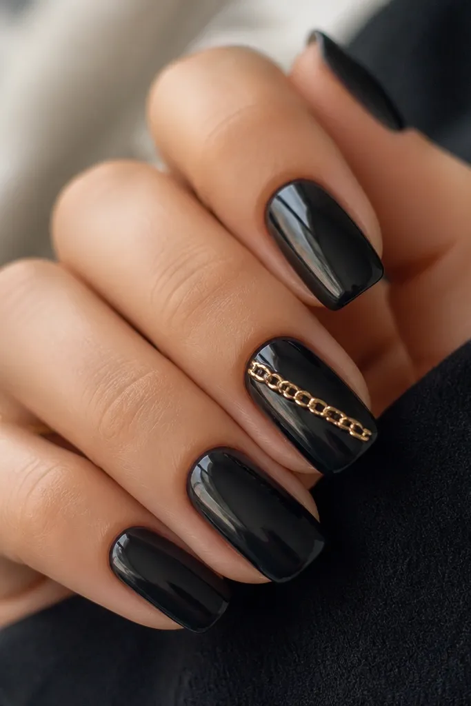 Glossy black nails with a single gold chain link line on one accent nail. The chain looks like tiny metal links placed diagonally from cuticle toward the side. Other nails stay plain black.