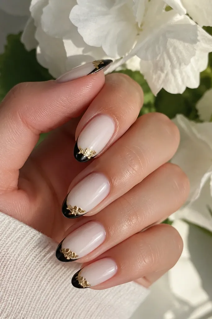 Short squoval nails with a sheer nude base, black only on the bottom half of the nail tip area, and a gold foil corner patch at the top edge of the black section.