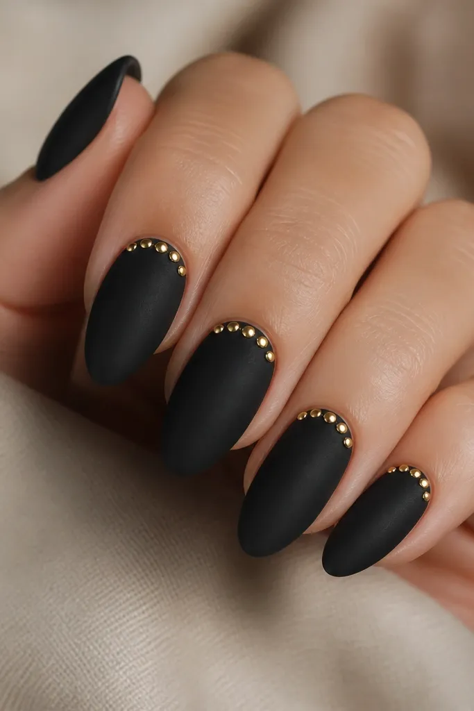 The nails are matte black. At the cuticle line, there are tiny gold studs or rhinestones arranged in a slight arc on each nail, with one or two studs on each side.