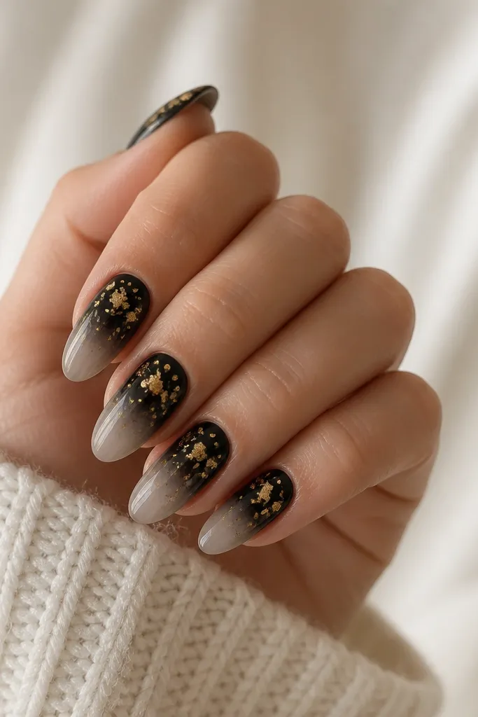 Nails start black at the cuticle and fade lighter toward the tips. Gold leaf flakes are scattered mainly on the upper half, with a few pieces near the center.