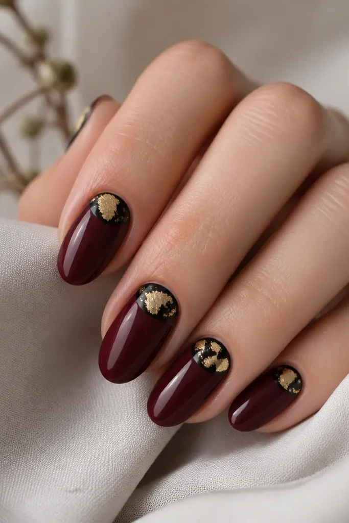Short almond nails with a glossy wine-red base, a crisp black half-moon near the cuticle, and irregular gold leaf pieces placed inside the half-moon area.