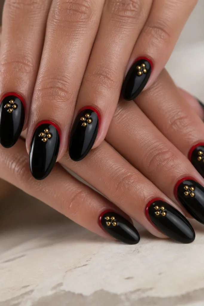 Glossy black nails with a red reverse French line at the cuticle, and three small gold studs in a triangle on the center of each nail.