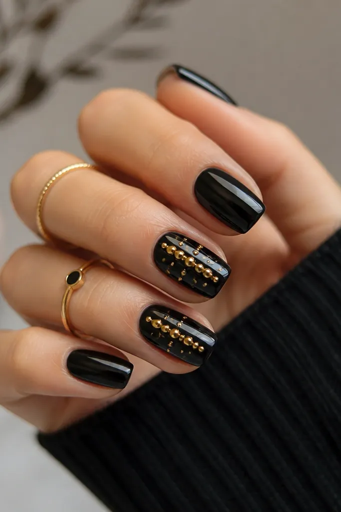 All nails are glossy black except two accent nails. On the accents, there are small gold studs clustered in a vertical line near the center with tiny gold rhinestones around them.