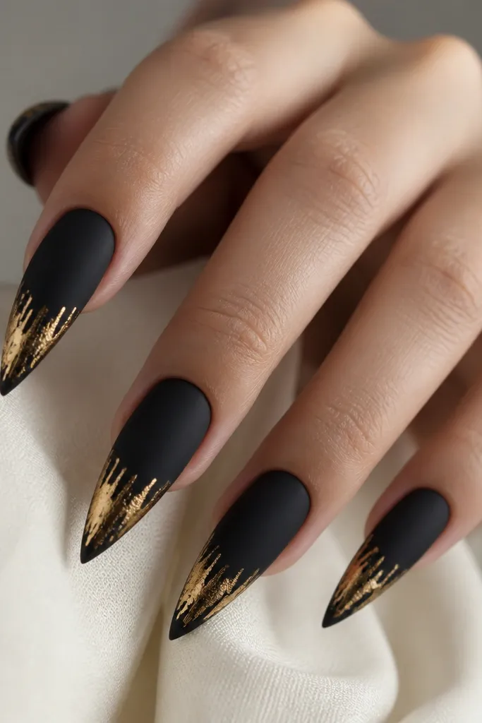 Stiletto nails coated in deep matte black with a soft "velvet" texture; each nail has a gold foil cluster at the tip with a downward drip effect and irregular edges.