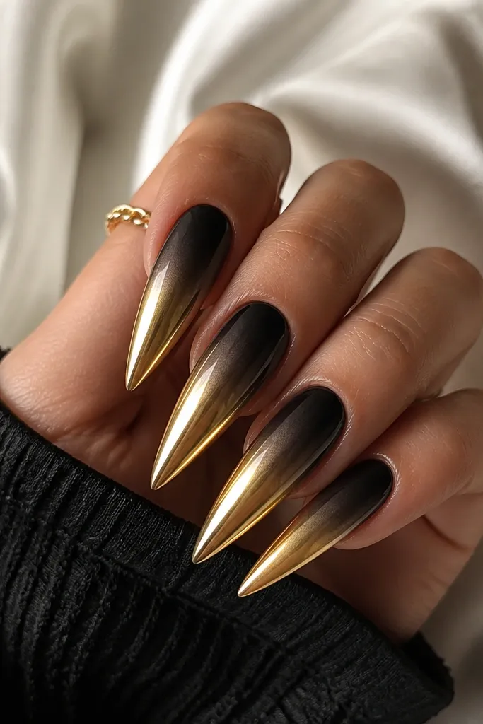 Stiletto nails with a smooth black-to-charcoal ombre that fades upward; the lower half transitions into a bright gold chrome tip with a sharp blend line.