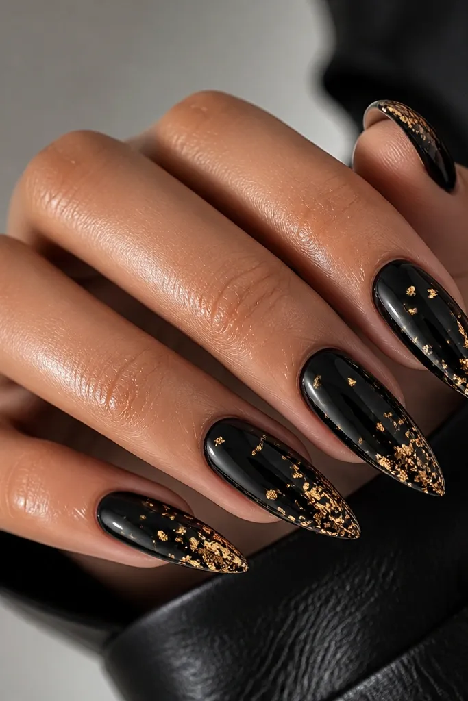 Glossy black stiletto nails with scattered small gold foil flakes across the top half and a heavier cluster near the tip.