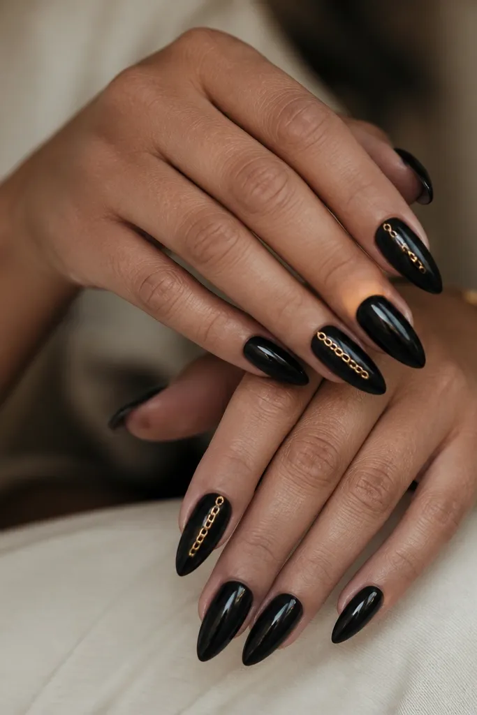 Black stiletto nails with a glossy finish; a thin gold chain link detail runs vertically down one nail per hand, with the rest in plain black and a few small gold link dots.