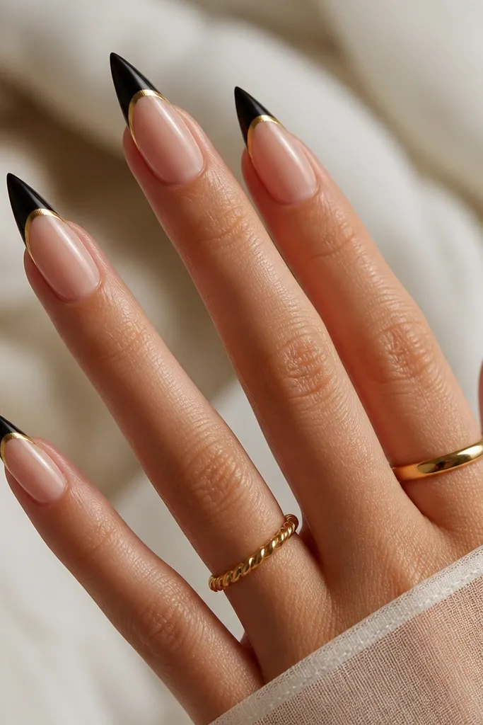 Stiletto nails with a black French tip line; the tip is outlined with a bright gold stripe following the smile curve; the base is sheer nude.
