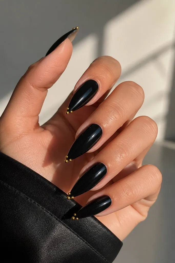Glossy black stiletto nails with two tiny gold studs placed at the tip corners like mirrored points; the rest is plain black.