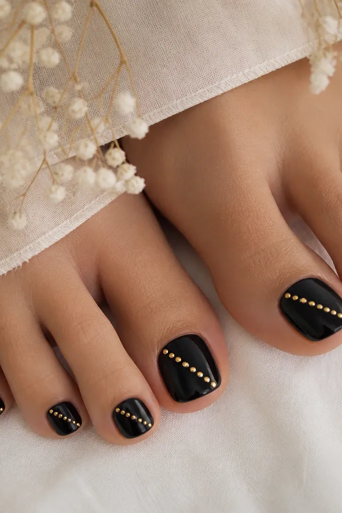 Toe nails painted glossy black. A straight line of tiny gold studs runs diagonally from near the cuticle to the outer tip.