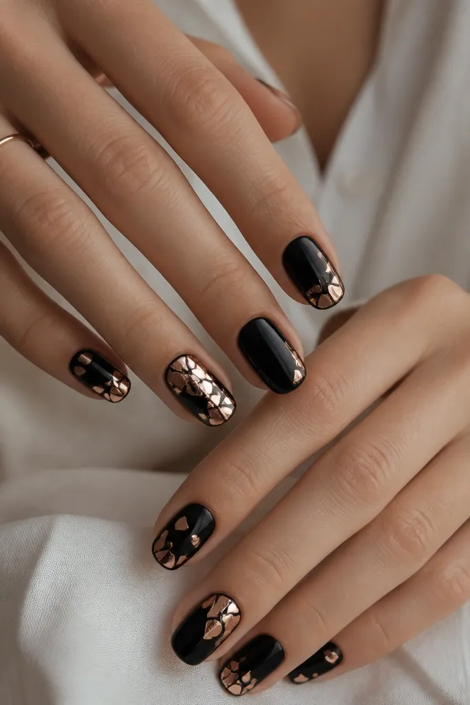 Black nails with scattered rose-gold mirror shards placed mostly near the corners and across one nail per hand. The mirror pieces look jagged and reflective.