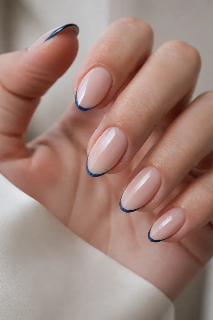 A set of medium almond nails showing a very thin cobalt blue French tip line. The blue looks glossy and mirror-like, with a cool silver reflection around it. The base is sheer nude, and the smile line is a smooth curve across each nail.