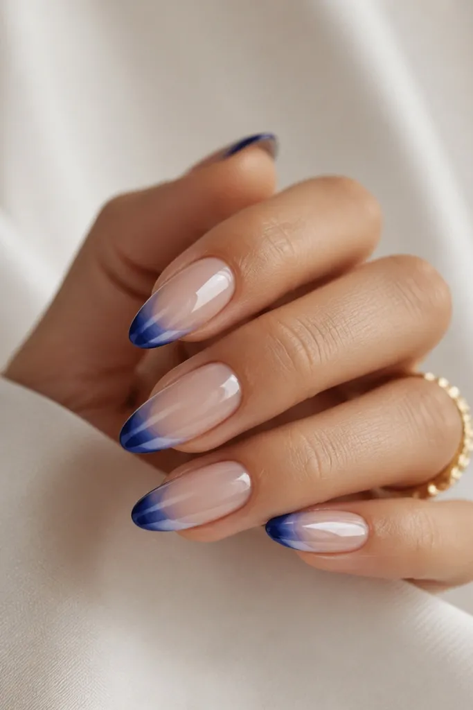 Almond nails showing a French tip that starts deep blue at the outer edge and fades into a lighter blue toward the center of the nail. The fade area has a mirror chrome finish. The base is sheer nude.
