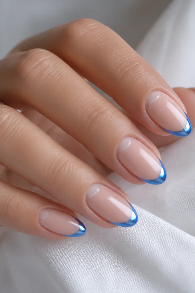 Short almond nails with a sheer nude base. At the cuticle area there is a clean negative space half-moon. The French tip is blue chrome with a bright mirror finish and a thin line.