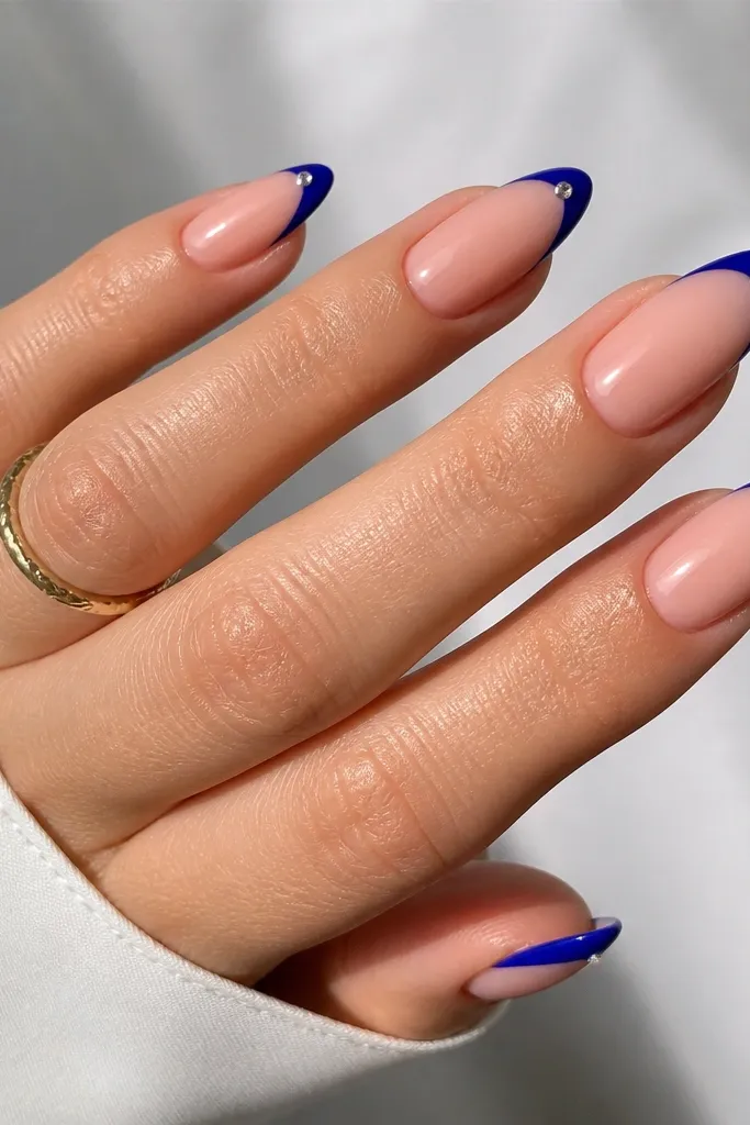 Almond nails with nude base and medium-width cobalt blue French tips; one rhinestone per nail at the outer corner of the tip; glossy finish.
