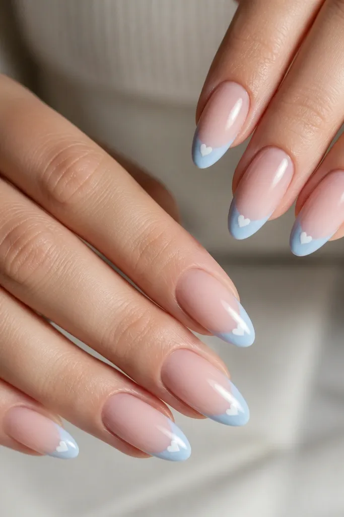 Almond nails with nude base; pale sky-blue French tips; a tiny white heart at the center of each blue tip; glossy finish.