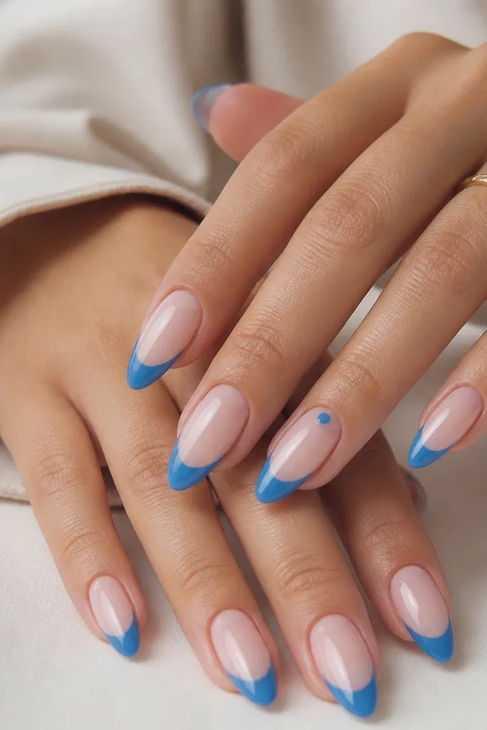 Almond nails in a soft natural nude base with a cerulean blue French tip that stops short of the sides. The cutout is a clean reverse half-moon at the base - nude is left exposed in a crescent shape, while the rest of the base is sealed. One accent nail has a tiny cerulean dot centered in the negative-space crescent, all with glossy top coat.