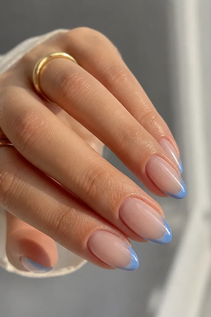 Short almond nails with a sheer nude base and glossy finish. The French tips are glazed blue with a slightly translucent center and darker blue edges for depth.