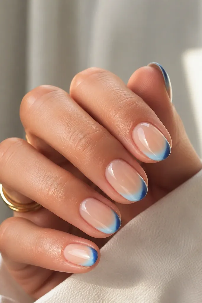 Short almond nails with a sheer nude base. French tips fade from lighter baby blue at the center to deeper blue at the outer edges.