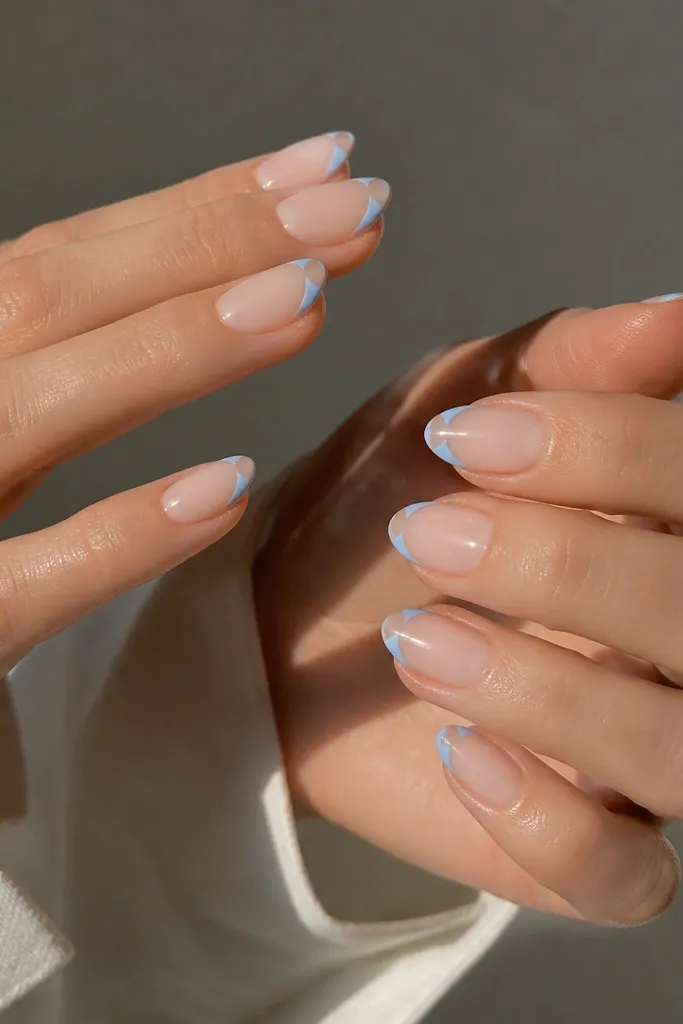 Short almond nails with sheer nude base. Powder blue French tips are painted around the sides and curve, leaving a small negative-space gap in the center of the tip.