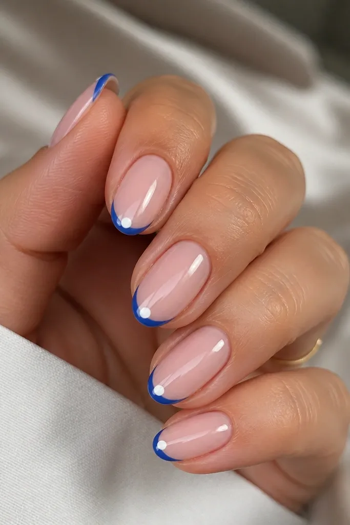 Short almond nails with sheer pink base. Blue French tips are narrow. Under the blue tip, there's a tiny white half-moon shape centered under the smile line.