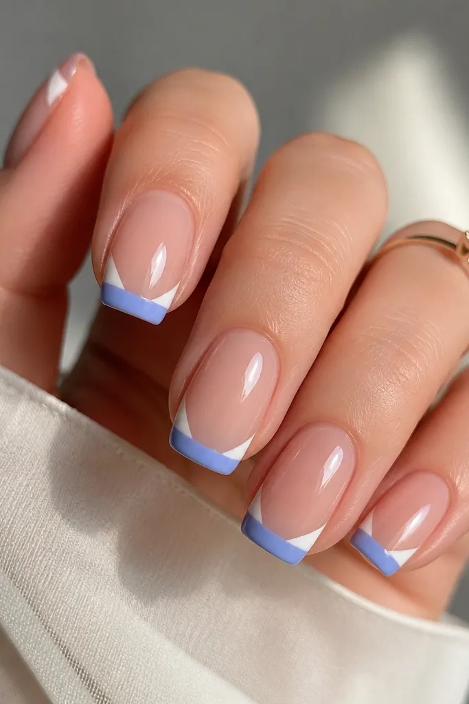 Medium square nails with a sheer nude base. The French tips are periwinkle blue with sharp V-shaped milky white corners at the side edges, like two tiny triangles. Everything is glossy with a smooth highlight.
