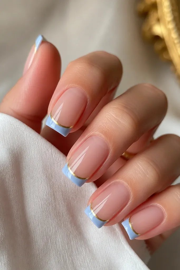 Square nails with a porcelain-blue French tip that is thin and crisp. A single thin gold pinstripe runs parallel to the smile line, just inside the blue. The cuticle area stays clear and glossy with a faint halo effect, so the blue starts slightly off the cuticle. The nails look clean, high-shine, and classy, with a glassy clear base and sharp edges.